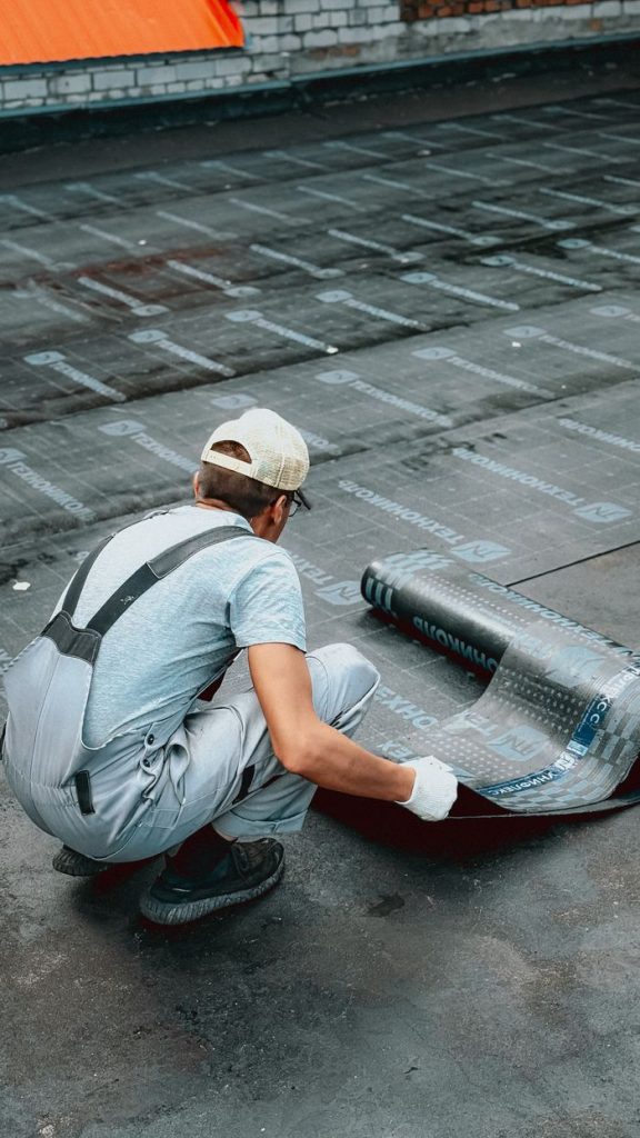 A worker kneels to lay a waterproofing membrane on a flat roof in an outdoor setting.